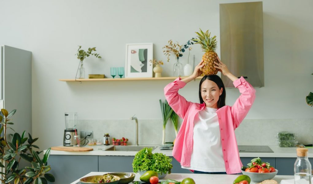 Asian woman in pink overshirt holding a pineapple in a modern kitchen with fresh produce and cheerful ambiance.