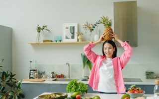 Asian woman in pink overshirt holding a pineapple in a modern kitchen with fresh produce and cheerful ambiance.