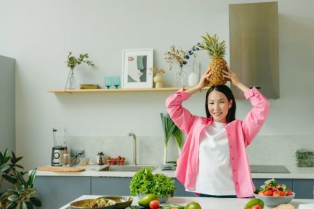 Asian woman in pink overshirt holding a pineapple in a modern kitchen with fresh produce and cheerful ambiance.